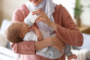 Muslim mother in hijab feeding her cute little baby with milk from bottle at home, closeup