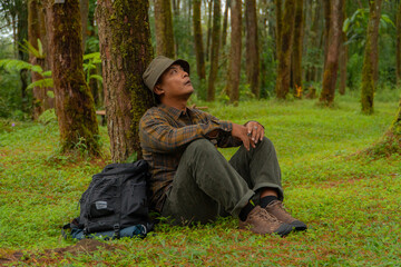 An adventurer is resting by leaning against a beautiful pine tree with a stretch of green grass. An adult man is resting while enjoying the tranquility of nature in a pine forest area