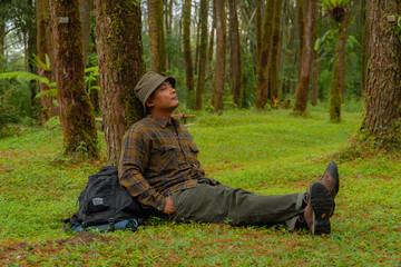 An adventurer is resting by leaning against a beautiful pine tree with a stretch of green grass. An adult man is resting while enjoying the tranquility of nature in a pine forest area