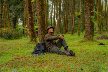 An adventurer is resting by leaning against a beautiful pine tree with a stretch of green grass. An adult man is resting while enjoying the tranquility of nature in a pine forest area