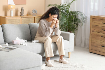 Young African-American woman sneezing in handkerchief and sitting on sofa at home. Allergy concept