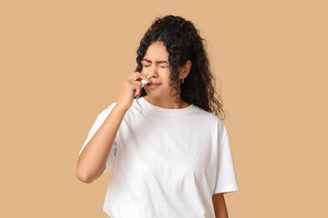Young African-American woman using nasal spray on beige background. Allergy concept