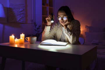 Young woman with flashlight reading book on table at home during blackout