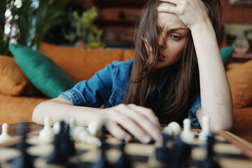 Stressed woman sitting on couch with head in hands contemplating chess moves on board in front of...