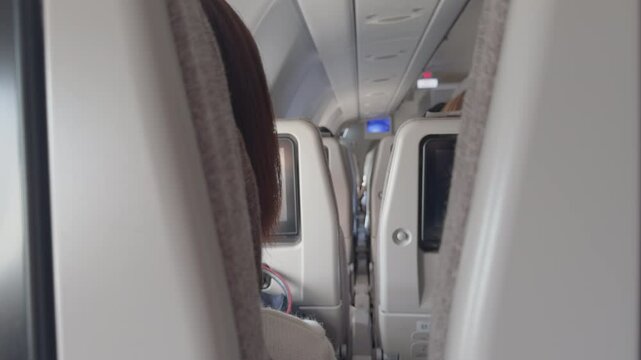 A unique perspective inside an airplane cabin, filmed between two seats, showing rows of passengers and a glimpse of two travelers communicating without revealing their faces.