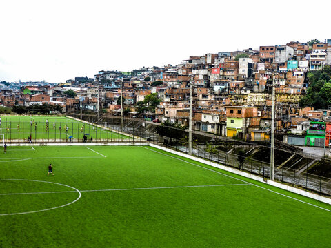 Campo de Futebol de V&aacute;rzea com Grama Sint&eacute;tica e Comunidade ao Fundo