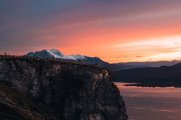 Fototapeta premium Cliff at Fjellheisen viewpoint in Tromsø, Norway, with a breathtaking sunset and snow-capped mountains in the background.