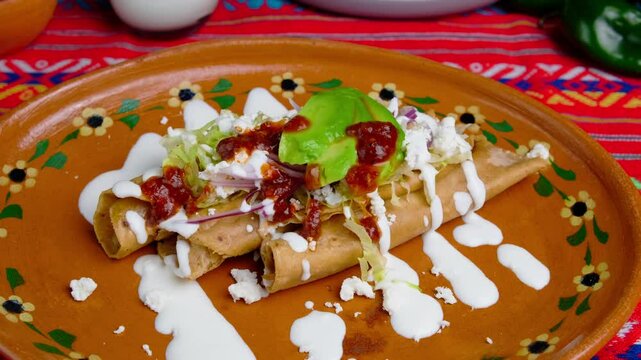 Taquitos garnished with lettuce, mexican cream, cheese, onion and avocado, served on a clay plate, mexican food