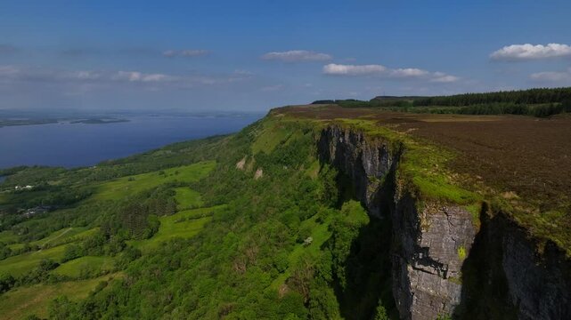 Cliffs of Magho, County Fermanagh, Northern Ireland, June 2023. Drone pushes forward in a sweeping aerial view of the craggy cliffside with woodland below and Lower Lough Erne in the distance.