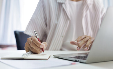 Fototapeta premium Asian man in casual clothes sitting at desk using laptop working and taking note, freelancer working online and doing part time job at home