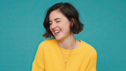 Fototapeta premium Young Woman Laughing with Eyes Closed, Wearing Vibrant Yellow Sweater, Solid Turquoise Background, Joyful Expression, Bright and Cheerful Mood, Colorful Contrast in Portrait