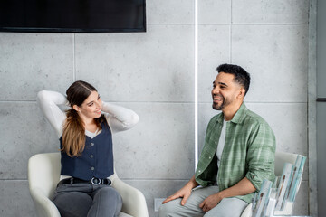Businesswoman and businessman are sitting in chairs in a waiting room, smiling and talking,...