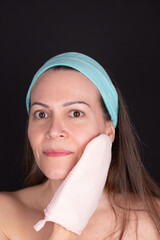 Woman in her 40's smiling and looking at camera with her hair up with a blue headband wipes her face with a light pink mitten. Black background. Concept of facial care and natural beauty