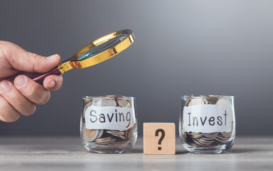 Man holding a magnifying glass near two glass jars filled with coins, one labeled Saving and the...