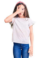 Fototapeta premium Cute hispanic child girl wearing casual white tshirt peeking in shock covering face and eyes with hand, looking through fingers with embarrassed expression.