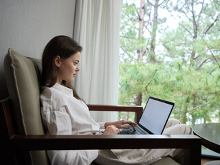 Young woman sitting comfortably in a modern armchair, focused on her laptop, surrounded by serene greenery, embodying relaxation and productivity in a bright workspace