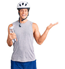 Young handsome man wearing bike helmet holding bottle of water celebrating victory with happy smile and winner expression with raised hands