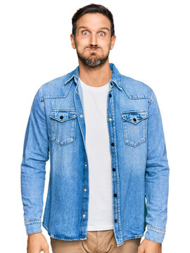 Young handsome man wearing casual denim jacket puffing cheeks with funny face. mouth inflated with air, crazy expression.