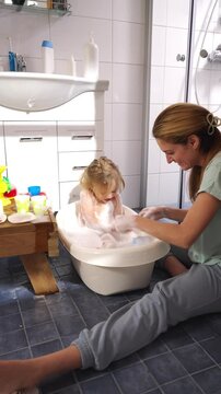 A cheerful mother and her toddler enjoy bath time in a bright bathroom. The child is sitting in a small bathtub, covered in soap bubbles, laughing joyfully. The mother, sitting on the tiled floor
