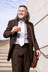 stylish mature businessman in classy suit with briefcase and takeaway coffee walking down urban stairs in city, concept of business and middle age man lifestyle