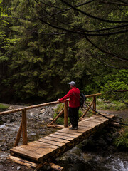 Fototapeta premium Man sitting on a bridge in the forest with a rocky path along a river. Man taking a break at the bridge by the river. Active vacation concept.