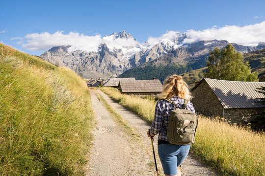 Woman hiking in Ecrins national park overlooking snowcapped Alps mountains with ancient stone houses along the path.