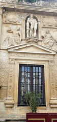 Fototapeta premium Stone facade with detailed carvings of figures and floral patterns; central statue depicting a muscular man, framed by two seated women, above a window with iron bars;