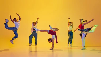 Dynamic image of stylish teen girls wearing casual attire, dancing hip hop, training against yellow background in neon light. Concept of modern dance, youth, active lifestyle, hobby