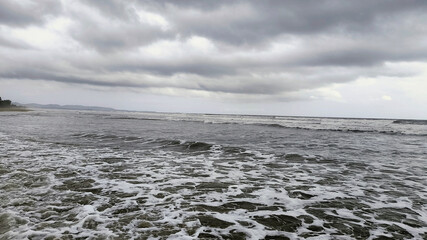 Dramatic waves crash along a shoreline under a cloudy, overcast sky. The dark tones of the sea and stormy atmosphere create a sense of power and nature's untamed beauty