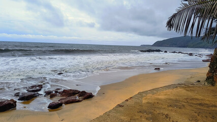 A serene beach with golden sand and rocky shore, set against a tropical backdrop and cloudy skies, evoking a peaceful and natural ambiance.