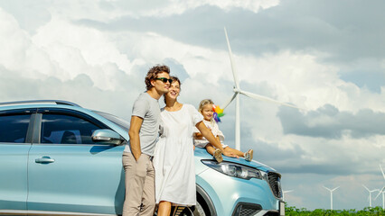 Concept of progressive happy family holding windmill toy and relax at wind farm with electric vehicle, happy family in the community with wind generators turbines, ev car