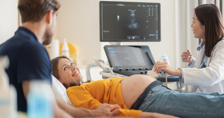 Obraz premium Asian Obstetrician Performs Ultrasound on Pregnant Patient, Explaining Baby Development. Modern Medical Equipment Displays Detailed Sonogram Images as Doctor Gestures Towards Screen