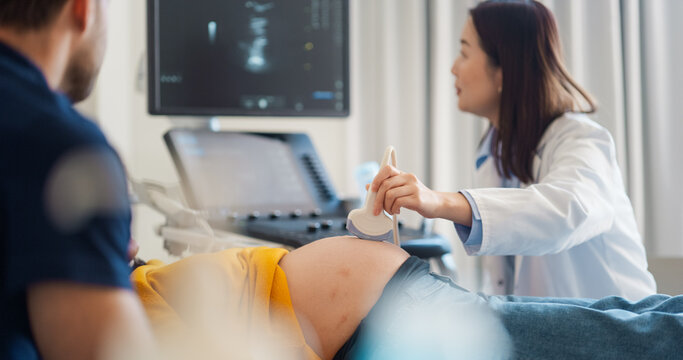 Caring Female Hospital Obstetrician Conducting an Ultrasound Exam for a Pregnant Woman. Young Couple Watching the Screen while Asian Doctor Describes the Baby’s Growth to Expectant Mother