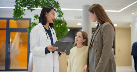 Asian Female Paediatrician Meeting a Little Girl with Mother in a Hospital Reception Hall. Doctor Using Tablet Computer to Show Medical Information and Test Results to a Young Family