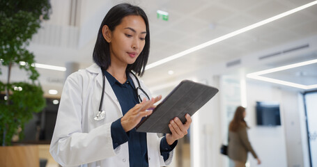 Portrait of an Asian Female General Practitioner Using Tablet Computer to as She is Browsing Insights About a Complex Medical Case. Healthcare Workers Walking Through a Modern Hospital Hall