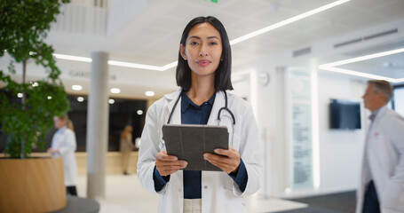 Fototapeta premium Portrait of a Female Doctor Browsing Information Online on a Digital Tablet Computer in a Modern Hospital Lobby. Asian Physician Looking at Camera, Posing. Colleagues and Patients Move in Background
