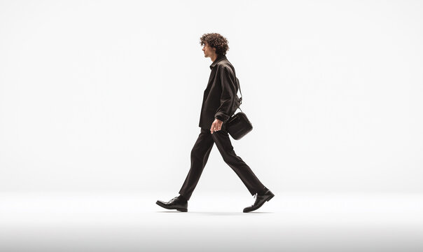 A man walks in the middle of an empty white space. He is wearing black trousers and a jacket with a shoulder bag. It is a cinematic, long shot.