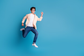 Obraz premium Joyful young man jumping enthusiastically against a vibrant blue background. Casual and stylishly dressed in trendy clothes.