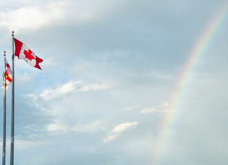 Two Canadian flags waving in the breeze against a cloudy sky, with a faint rainbow emerging in the...