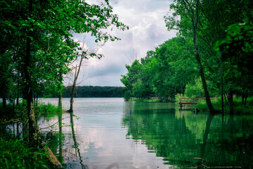 Obraz premium A serene lakeside scene surrounded by lush green trees, reflecting in calm water under a cloudy sky, creating a peaceful and natural landscape.