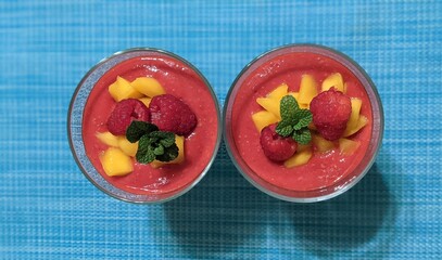 Top-down view of two raspberry and mango desserts in glass cups, garnished with fresh raspberries, diced mango, and mint leaves on a bright blue textured background.