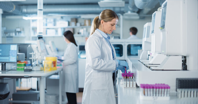 Team of Biomedical Engineers Work in a Lab. Senior Researcher Adding Samples to Test Machine, Asian Female Adjusting Settings on a Digital Microscope while Male Colleague Carrying Medical Glassware - Powered by Adobe