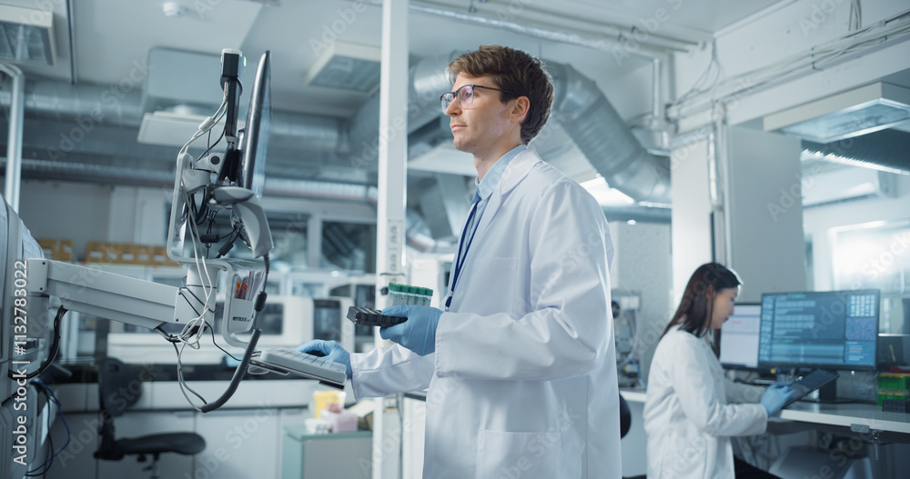 Poster portrait of a young male scientist holding a set of test tubes and using a computer while standing i