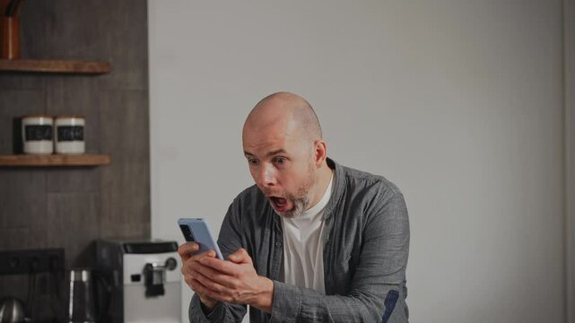 A middle-aged man is in his kitchen when he suddenly receives great news on his smartphone. The expressive man is overjoyed, displaying bright emotions of happiness and excitement.