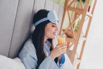 Charming woman enjoying a fresh morning with juice in bed wearing blue sleepwear and eye mask