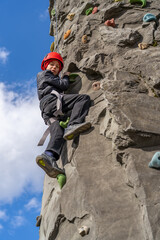 A child in a safety helmet climbs a rock wall at an outdoor adventure park on a sunny day,...