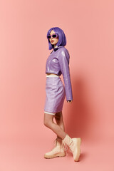 Beautiful woman with purple hair and stylish outfit poses confidently against a pink backdrop.