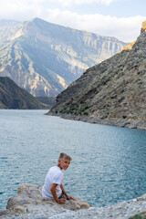 Fototapeta premium A young boy sits on a rocky shore, gazing at the tranquil lake surrounded by towering mountains in the warm light of late afternoon
