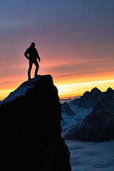 Obraz premium Silhouette of a man standing on the edge of a mountain cliff on the background of sunset nature and the sky, creatine concept of achieving a goal, success