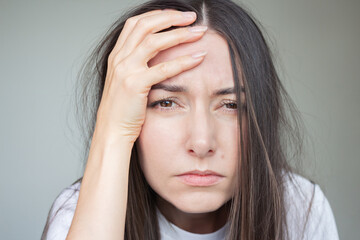 Fototapeta premium A young Caucasian brunette woman in white clothes is shaggy and sleepy, suffering from a headache, looking sadly at the camera, holding her head with her hand.